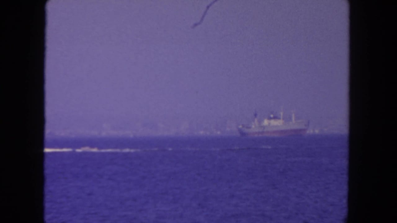 Distant ship on a blue ocean beneath a hazy sky, framed by dark film borders.