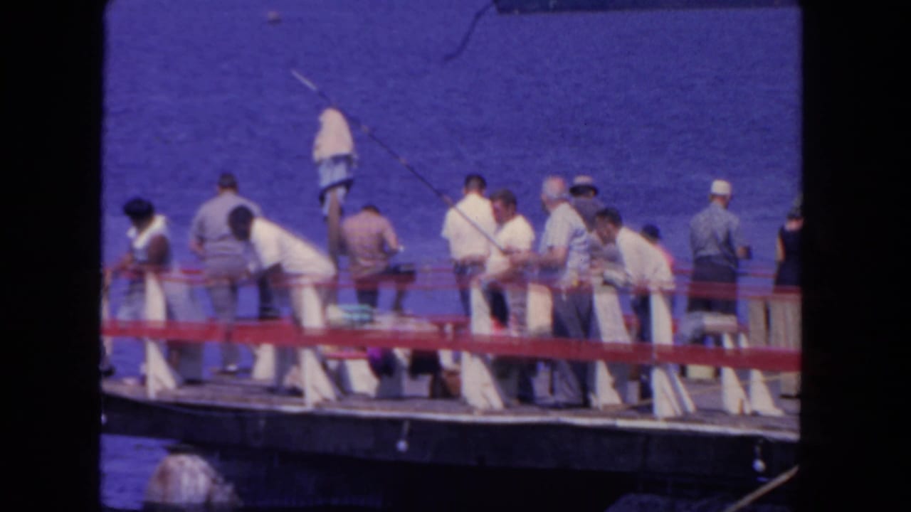 1962 San Diego 8mm frame showing men in light clothing and hats fishing from a wooden pier above blue water.