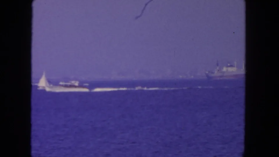1962 coastal home-movie frame with a sailboat, powerboat wake, and larger vessel in hazy water off San Diego.