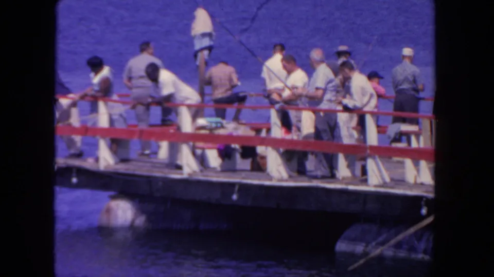 1962 San Diego home-movie frame showing men with fishing rods along a red-railed wooden pier above blue harbor water.
