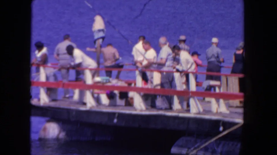 Men in casual summer clothing fish from a red-railed San Diego harbor pier in this 1962 8mm frame.