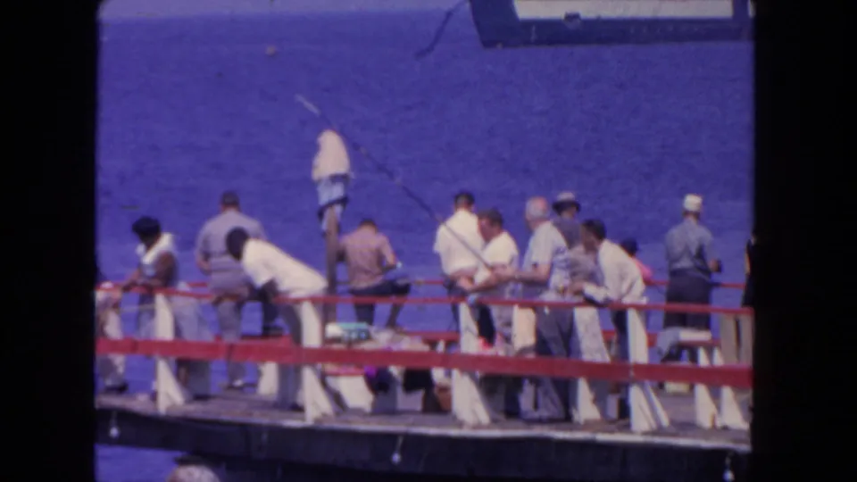 Final 1962 San Diego frame of men fishing and sitting along a red-railed pier over deep blue harbor water.