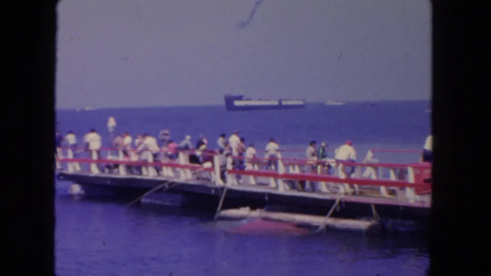 1962 San Diego pier scene with people in light clothing fishing and looking toward the water as a cargo ship sits on the horizon.