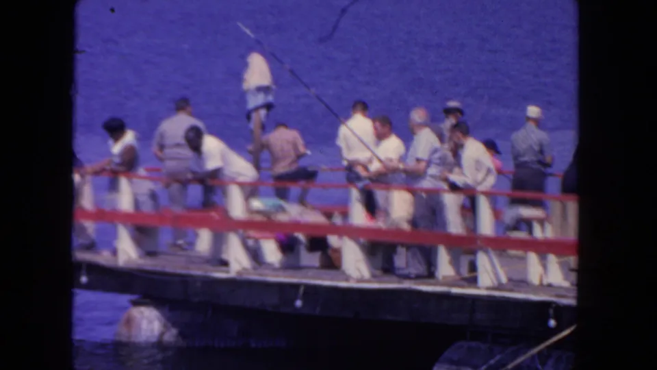 1962 San Diego home-movie frame showing a closer cluster of adults along the pier railing with fishing rods angled over the harbor.