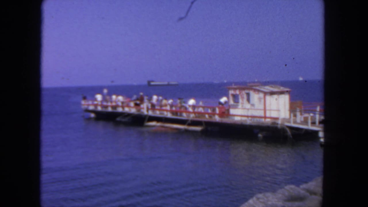 1962 San Diego home-movie frame showing people gathered on a seaside pier over calm water, with a large ship offshore.