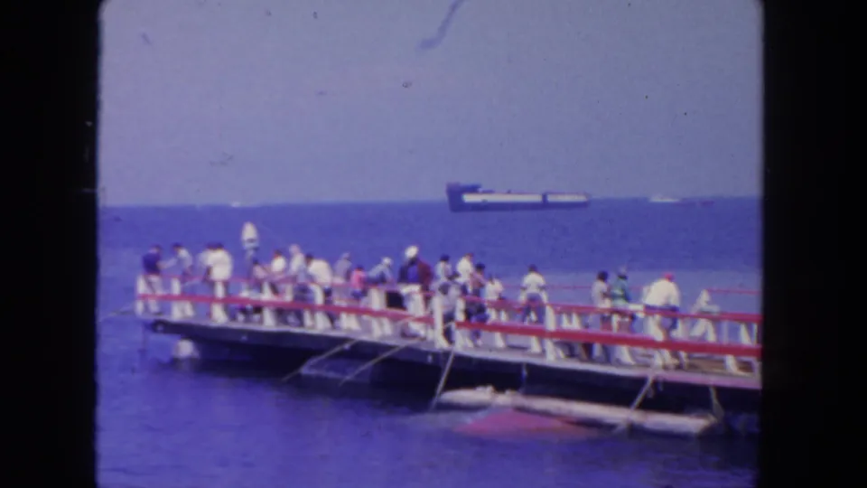 1962 San Diego pier scene with a crowd along the railing, apparent fishing activity, blue water, and a cargo ship on the horizon.