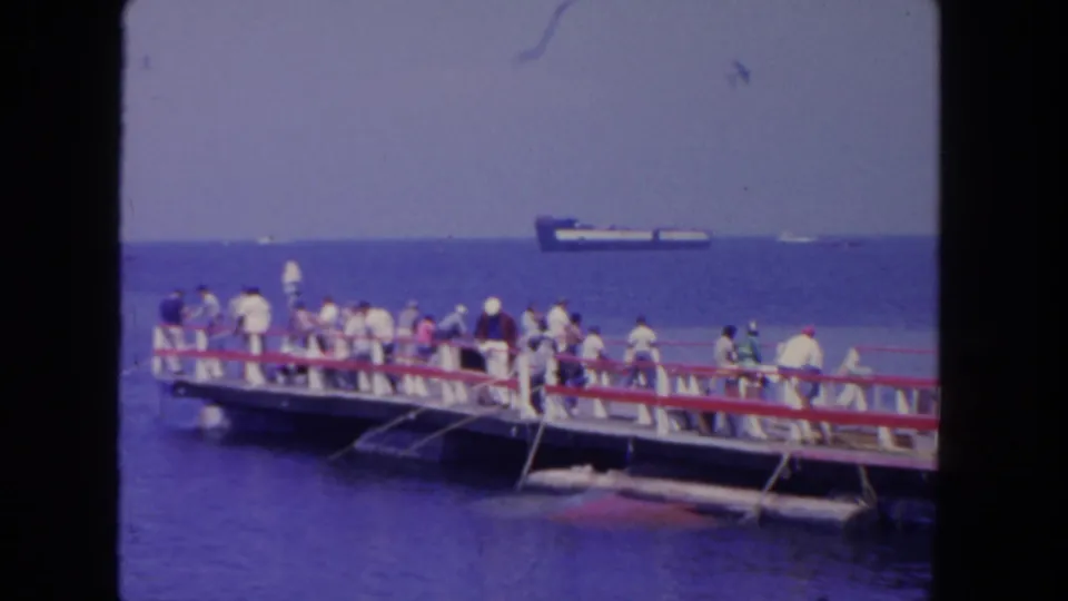 1962 San Diego home-movie frame of people on a pier, apparent fishing lines over the water, and an offshore ship beyond the railings.