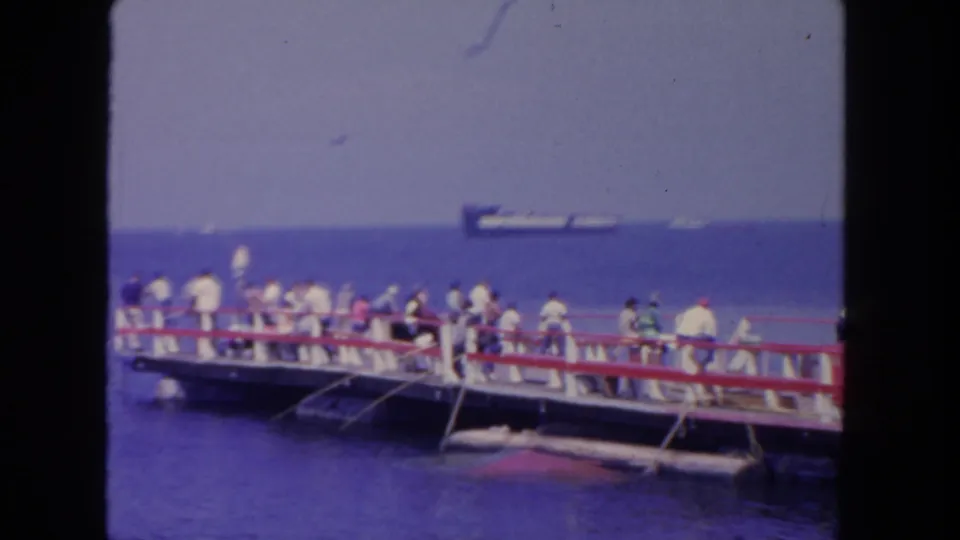 1962 San Diego pier footage frame showing a public seaside crowd above blue water, with a ship offshore and soft 8mm color.