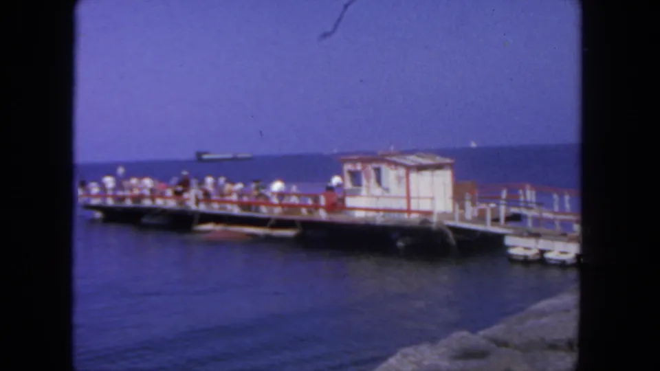 1962 San Diego waterfront frame with people lining a small pier, a red-and-white hut at one end, open water, and a distant ship.