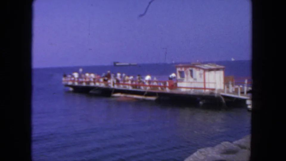 1962 San Diego 8mm frame showing the pier shifted in the pan, with visitors, a small hut, rocky foreground, and ship traffic offshore.