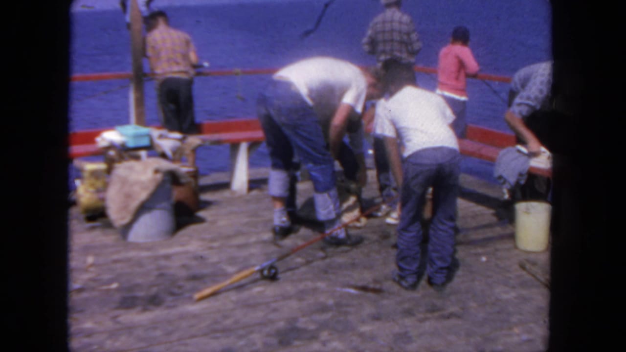 People fishing and standing on a wooden pier beside blue water.