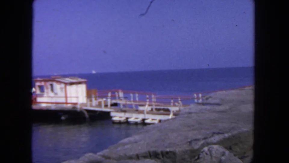 1962 San Diego waterfront view of a small white shack with red trim on a pier, with blue water and rocky shoreline nearby.