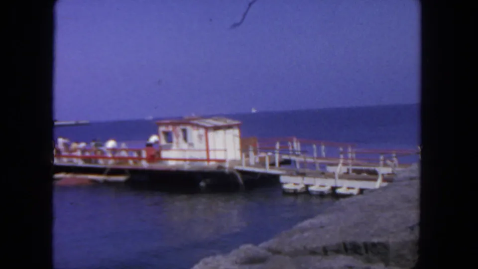 1962 San Diego waterfront frame with people near a red-trimmed pier shack, small floats, blue water, and rocks in the foreground.