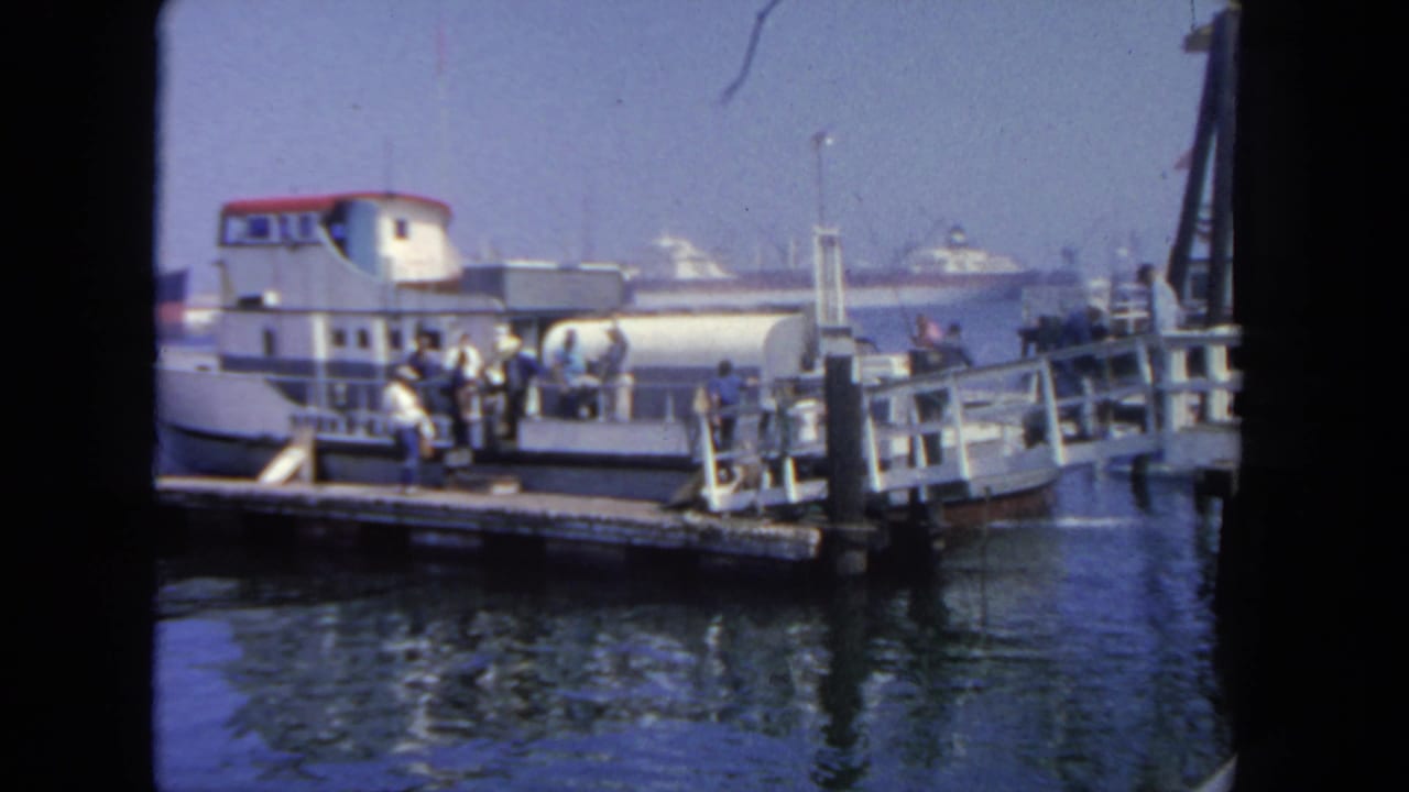 Passengers stand on a wooden dock beside a white-and-red passenger boat in a harbor.