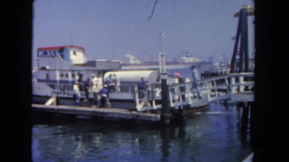 Wide 1962 San Diego harbor frame showing a passenger boat at the dock with people nearby and ships in the distance.