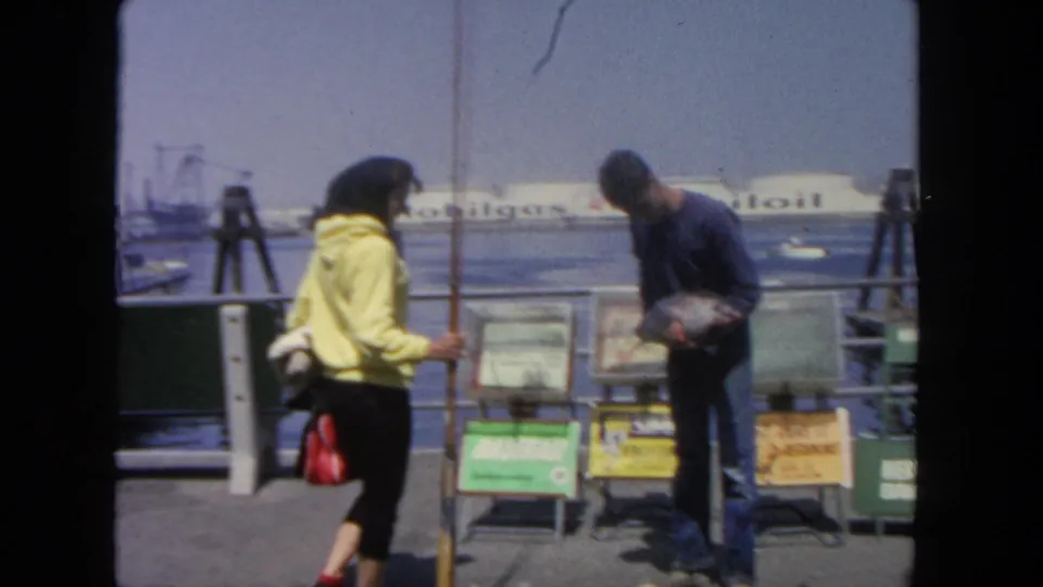 At the San Diego harbor rail, a woman in a yellow top and a man holding a fish stand beside posted informational signs.