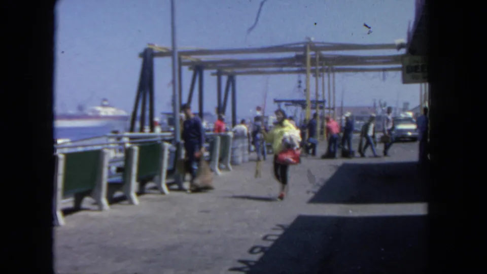 1962 San Diego harbor pier scene with people carrying bags near railings and a ferry boarding structure.