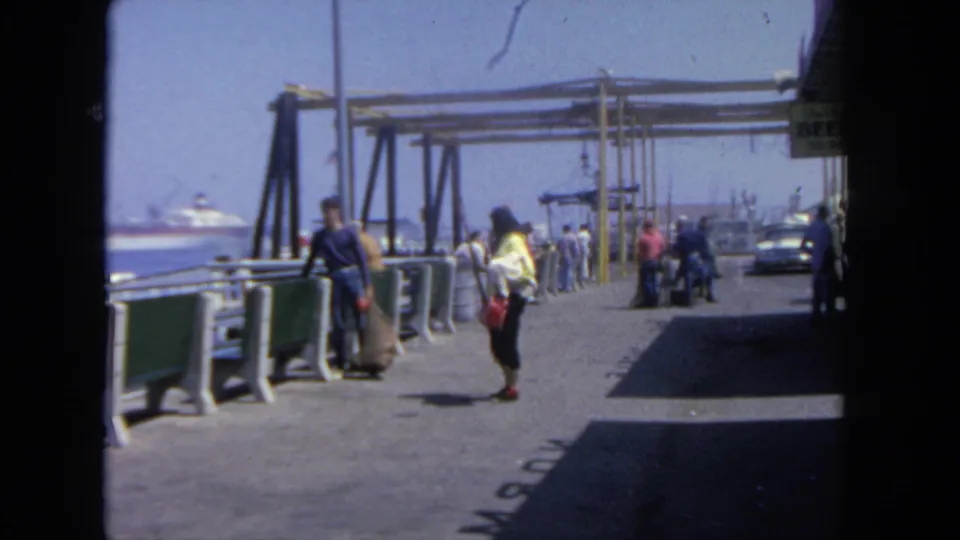 1962 San Diego pier view with visitors and passengers near a waterfront boarding area and distant harbor ships.