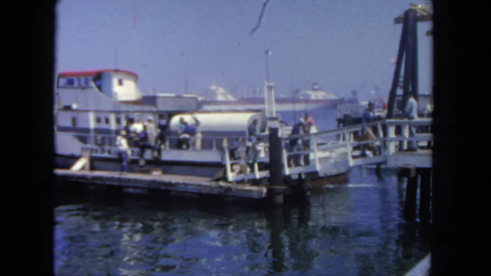 1962 San Diego ferry landing with a small ferry, gangway, waiting passengers, and harbor vessels beyond.