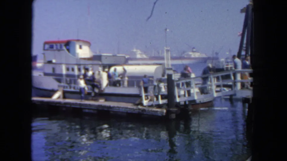 1962 San Diego harbor ferry boarding view with people at the gangway and larger vessels in the distance.
