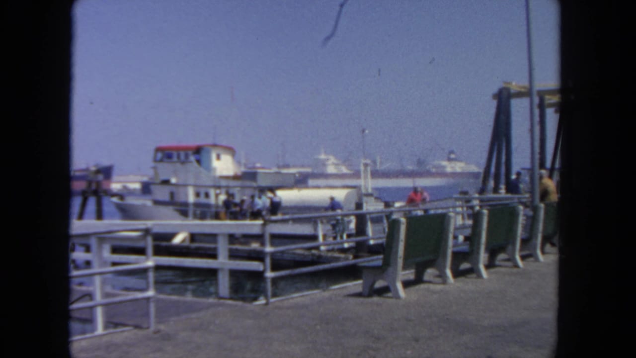 Ferry dock with passengers near railings, benches, water, and distant boats.