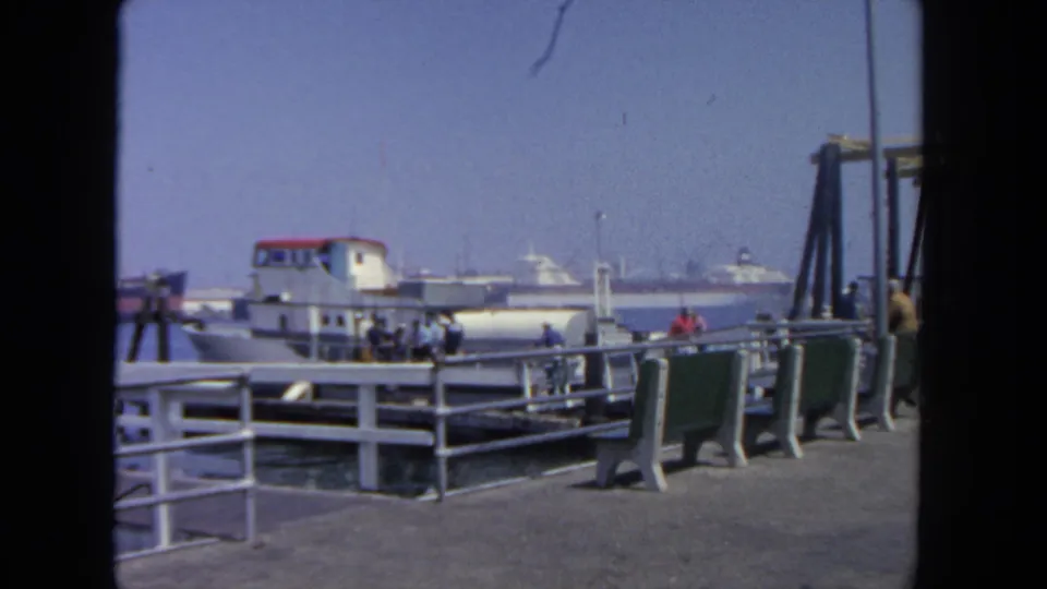 1962 San Diego harbor pier view with a white ferry, red roof, green benches, railings, pedestrians, and boats in the distance.