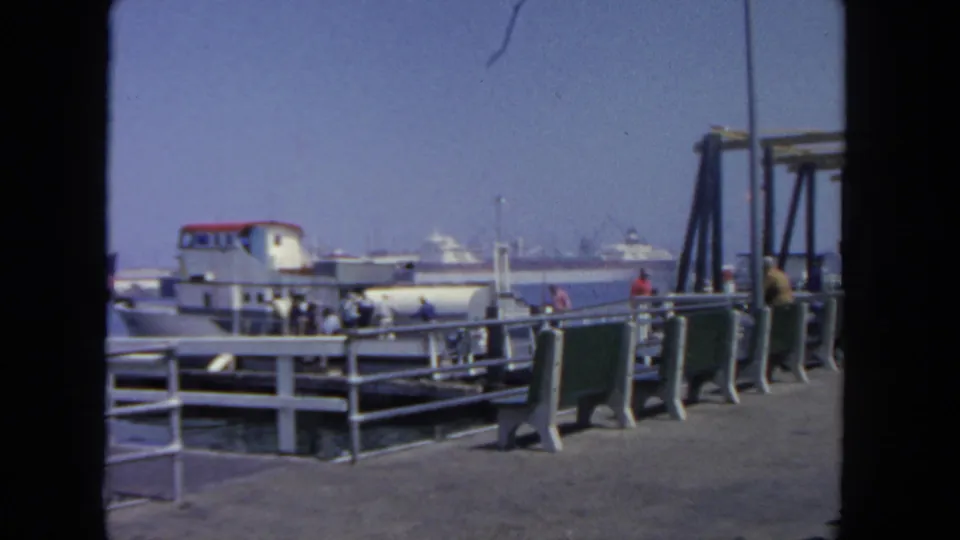 1962 San Diego pier scene showing pedestrians by a moored white ferry, green benches, railings, and distant harbor boats.