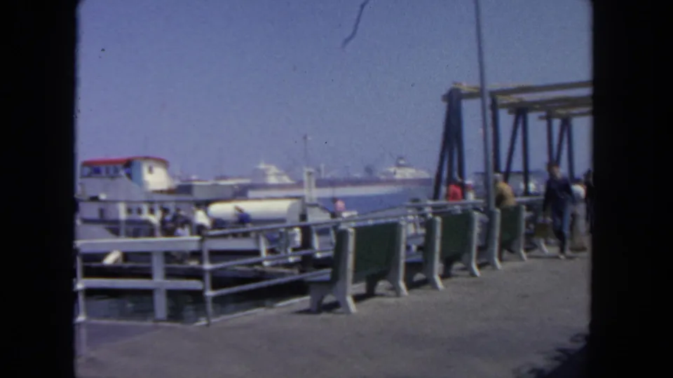 1962 San Diego waterfront view with passengers near a ferry, a row of green benches, white railings, and harbor boats beyond.
