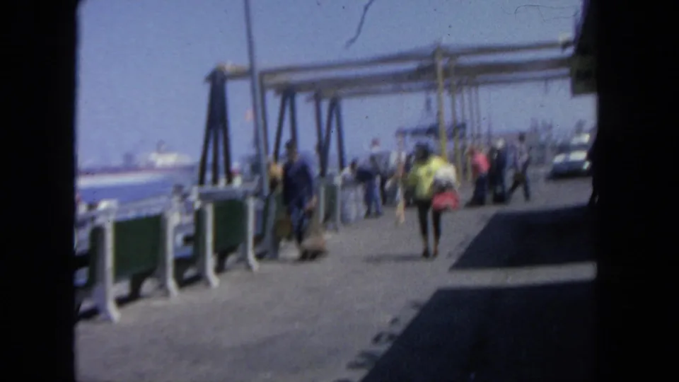 1962 San Diego pier walkway with a small crowd, green benches, railings, overhead dock structure, and boats on the harbor.