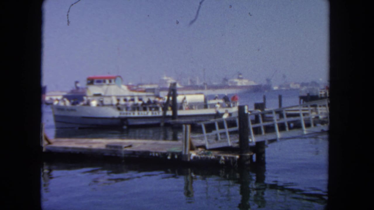 1962 San Diego harbor view with a white ferry at a pier, a sloped dock ramp, calm water, and ships in the distance.