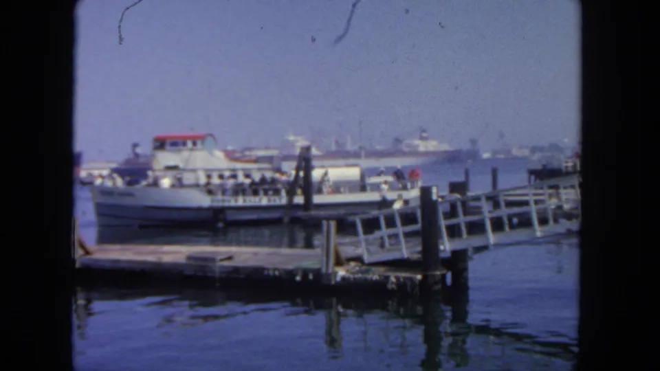 1962 San Diego harbor view with a white ferry at a pier, a sloped dock ramp, calm water, and ships in the distance.