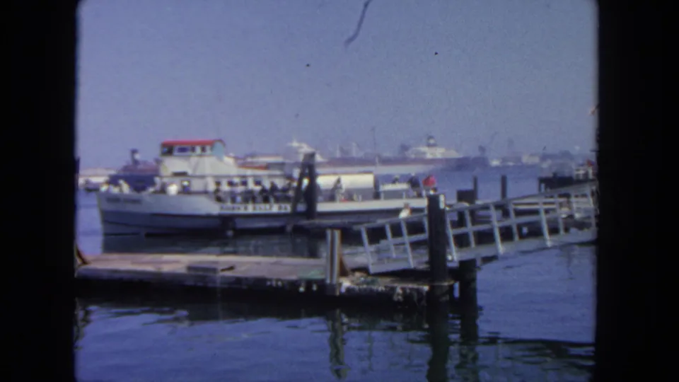 The ferry remains tied at a San Diego pier, with the ramp and mooring posts foregrounded against hazy harbor traffic.