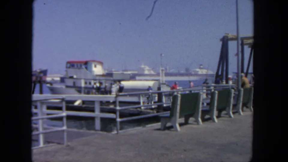 Several passengers walk away from the ferry along the San Diego dock, framed by railings, pier posts, and distant harbor vessels.