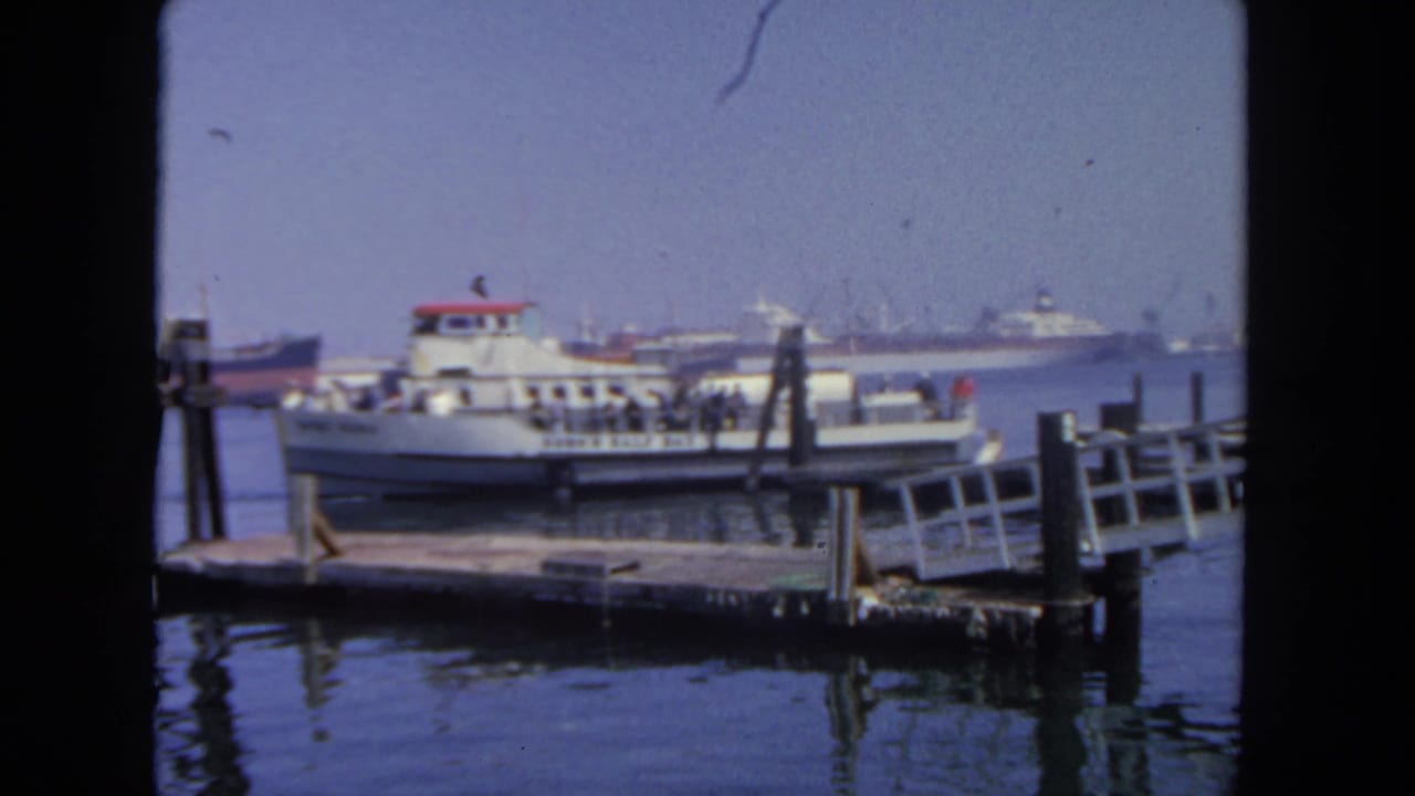 White passenger ferry beside a wooden pier with people on deck and harbor ships in the distance.