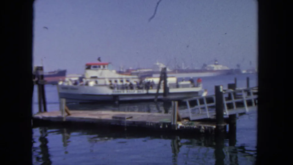 1962 San Diego home-movie frame showing a small passenger ferry at a wooden pier, with passengers aboard and larger harbor vessels behind it.