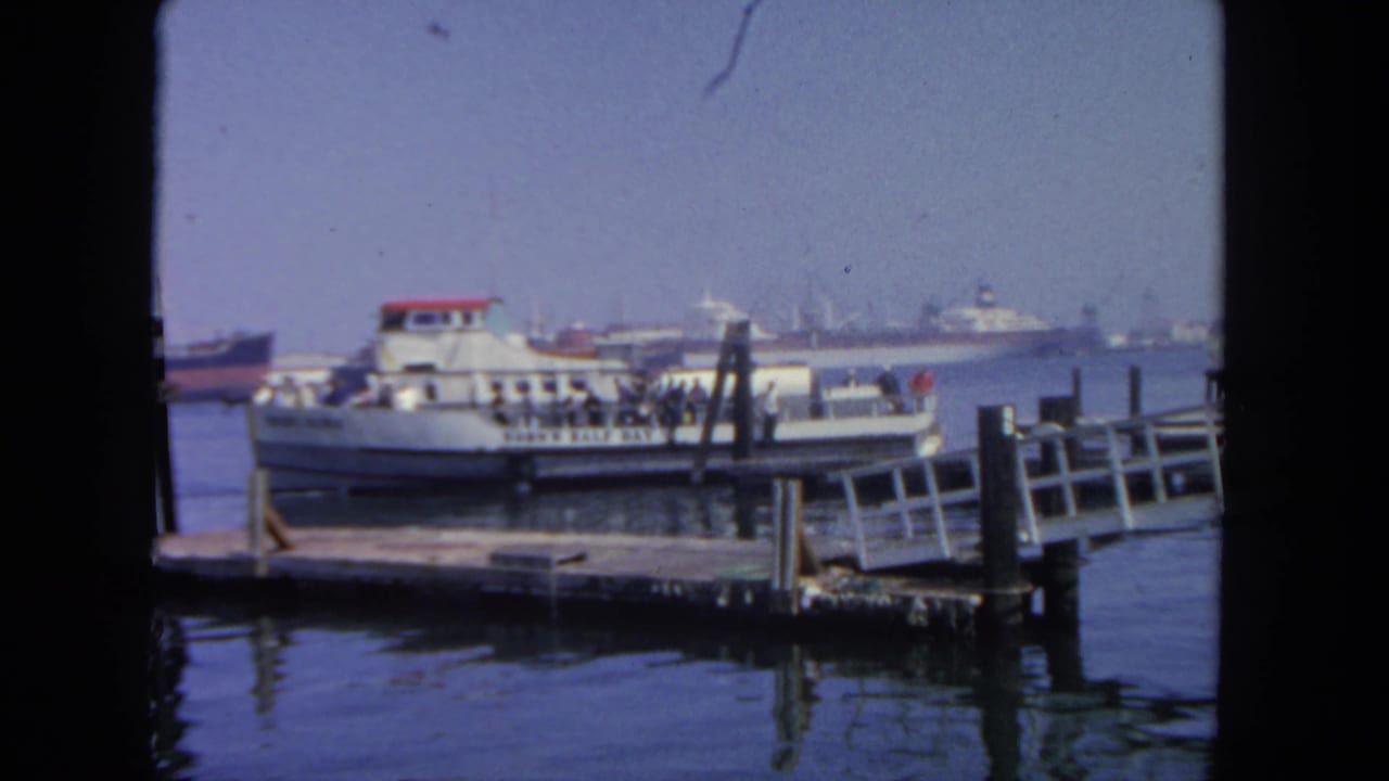 Passenger ferry moored near a wooden pier, with deck passengers and distant ships across the harbor.