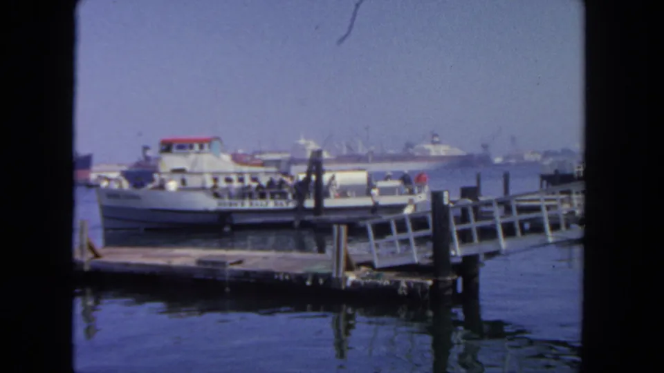 1962 San Diego harbor frame showing a ferry at a wooden pier, deck passengers, dock railings, and large ships softened by haze in the distance.