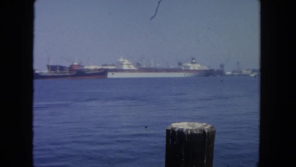 1962 San Diego harbor view with blue water, distant cargo ships, a mooring post, and dark 8mm frame borders.