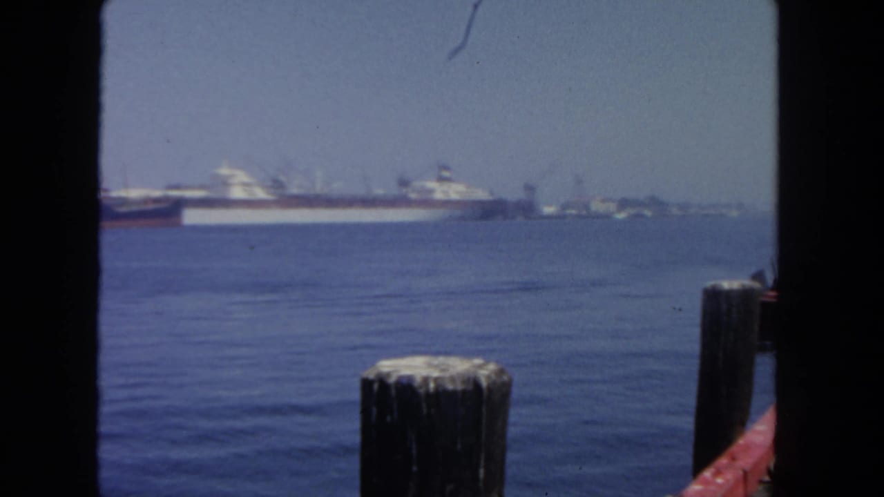 San Diego harbor in 1962, with cargo vessels on the horizon, calm water, a foreground dock post, and visible 8mm gate edges.