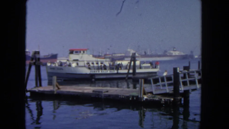 A small ferry at a San Diego dock in 1962 carries visible passengers, with wooden pier structures and cargo ships behind it.