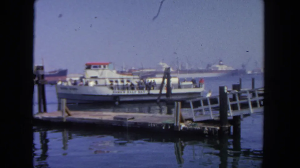 1962 San Diego dock scene with a small passenger ferry, visible riders, pier railings, and cargo vessels across the harbor.