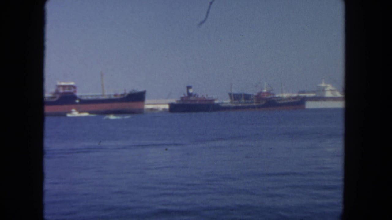 1962 San Diego harbor view with distant cargo ships or tankers, calm blue water, and a small motorboat near the foreground.