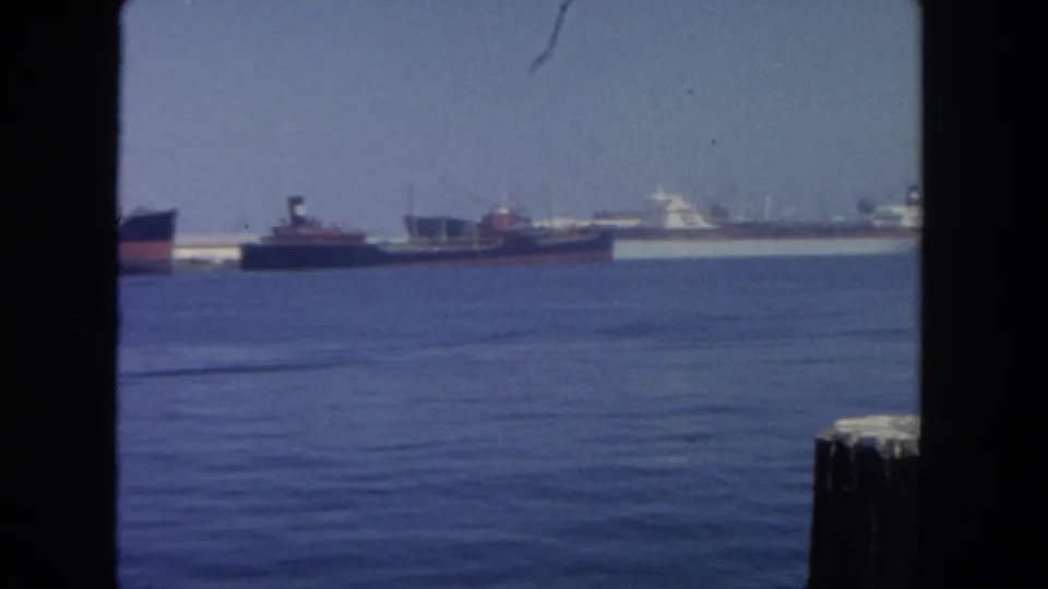 1962 harbor view in San Diego with distant ships, calm water, dark framing, and a pier piling visible at the lower right.