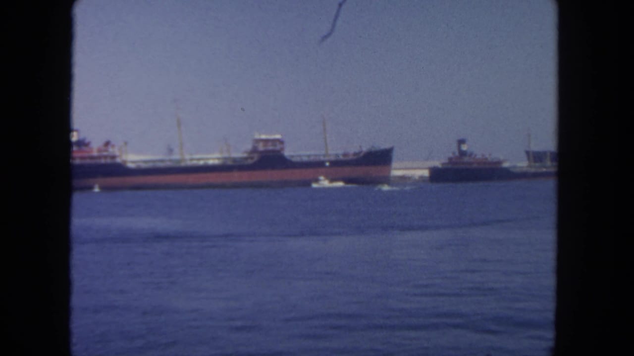 1962 San Diego harbor scene showing a large red-and-dark-hulled vessel, smaller tugboats, calm water, and distant pier structures.