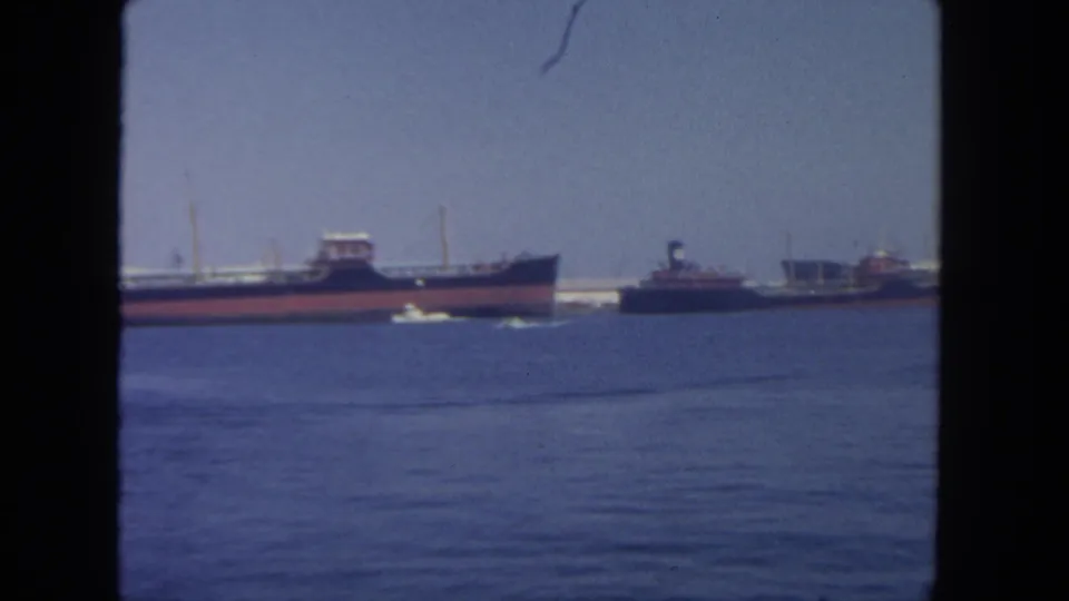 Soft 8mm view of San Diego harbor in 1962 with a cargo ship, tugboat, small white boat wake, and dock structures in the distance.