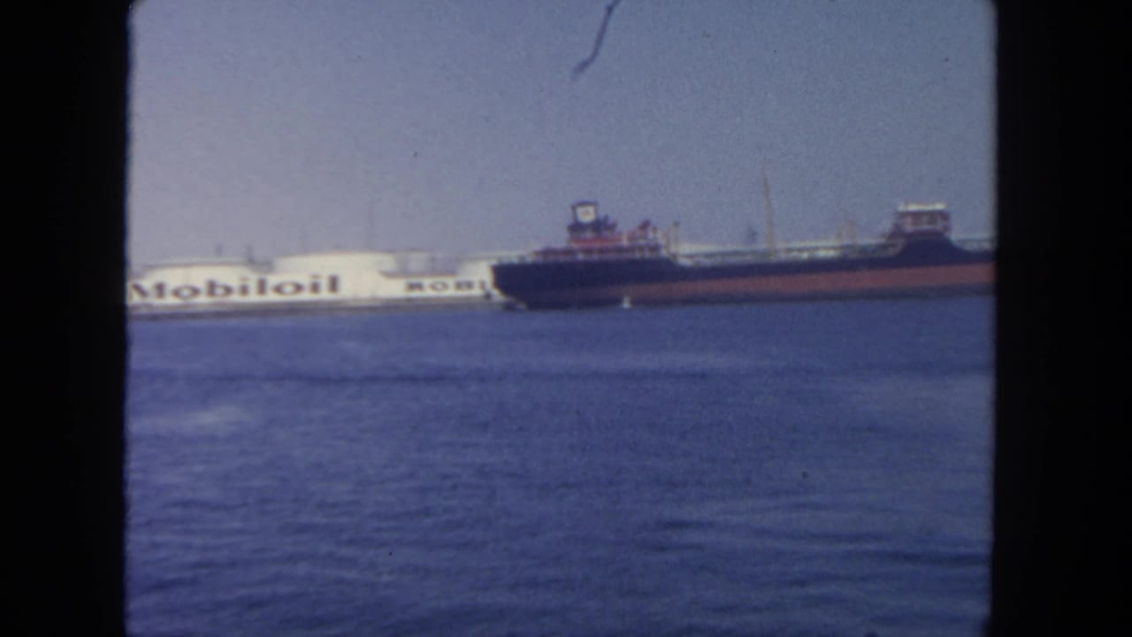 1962 San Diego harbor view with a dark cargo ship crossing in front of a white Mobil oil tanker across calm blue water.