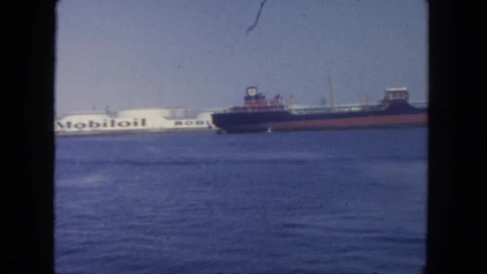 1962 San Diego harbor view with a dark cargo ship crossing in front of a white Mobil oil tanker across calm blue water.