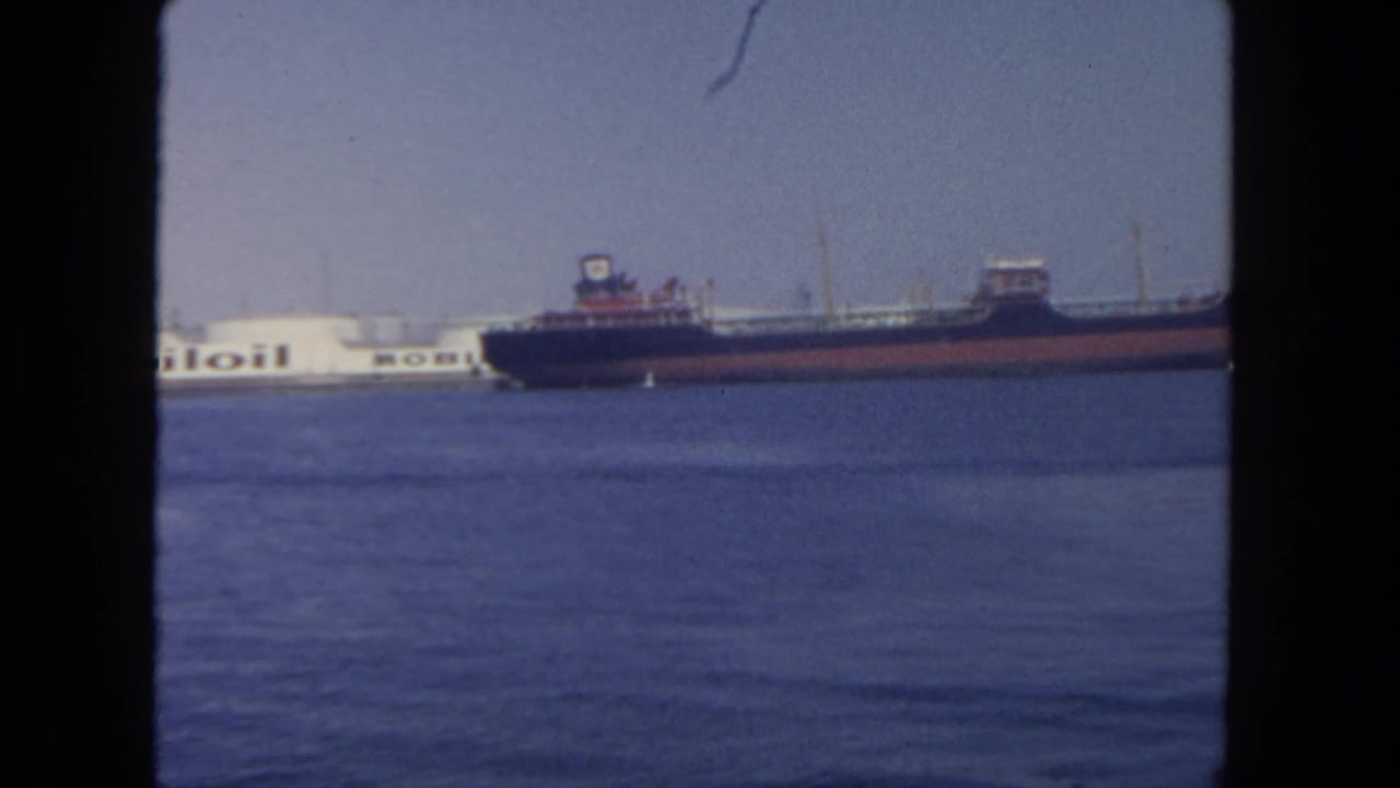 Distant 1962 San Diego harbor frame showing a cargo ship beside the visible Mobil tanker lettering, with open water in the foreground.