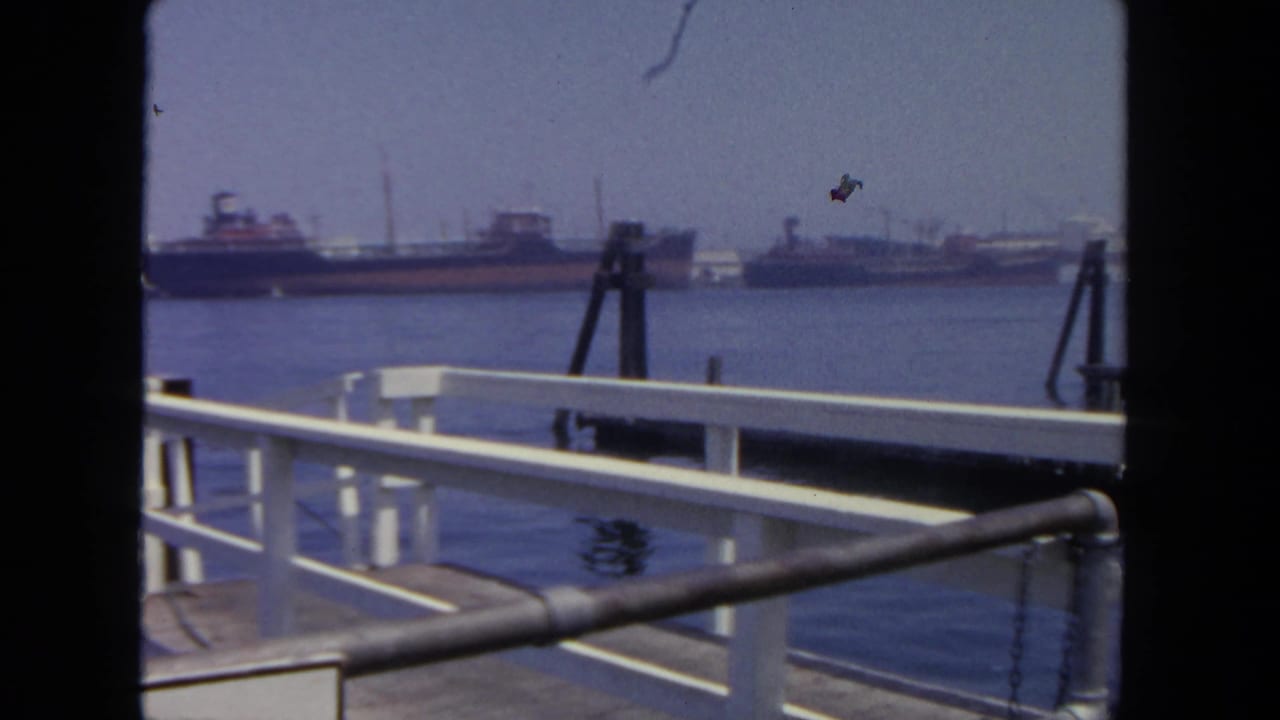 1962 San Diego harbor view from a dock, with white railings in the foreground and several distant tankers on calm water.