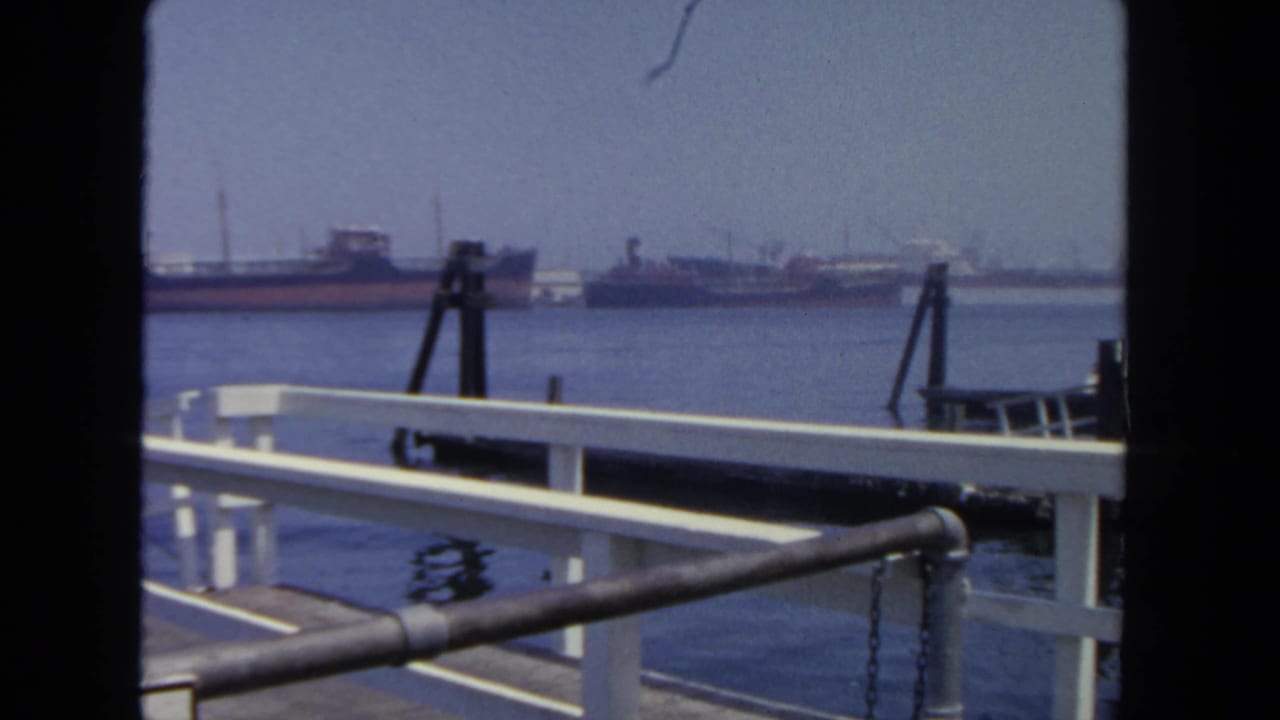 Dockside view in San Diego harbor, with white railings, a dark foreground rail, calm water, and tankers along the far shore.